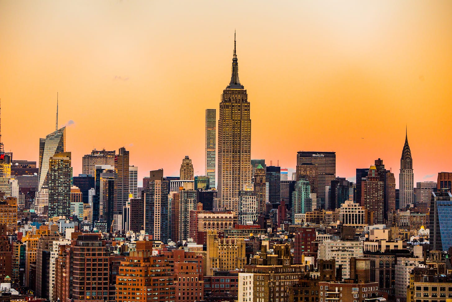 The New York City skyline amidst an orange sky. The Empire State Building rises prominently in the middle of the photo.