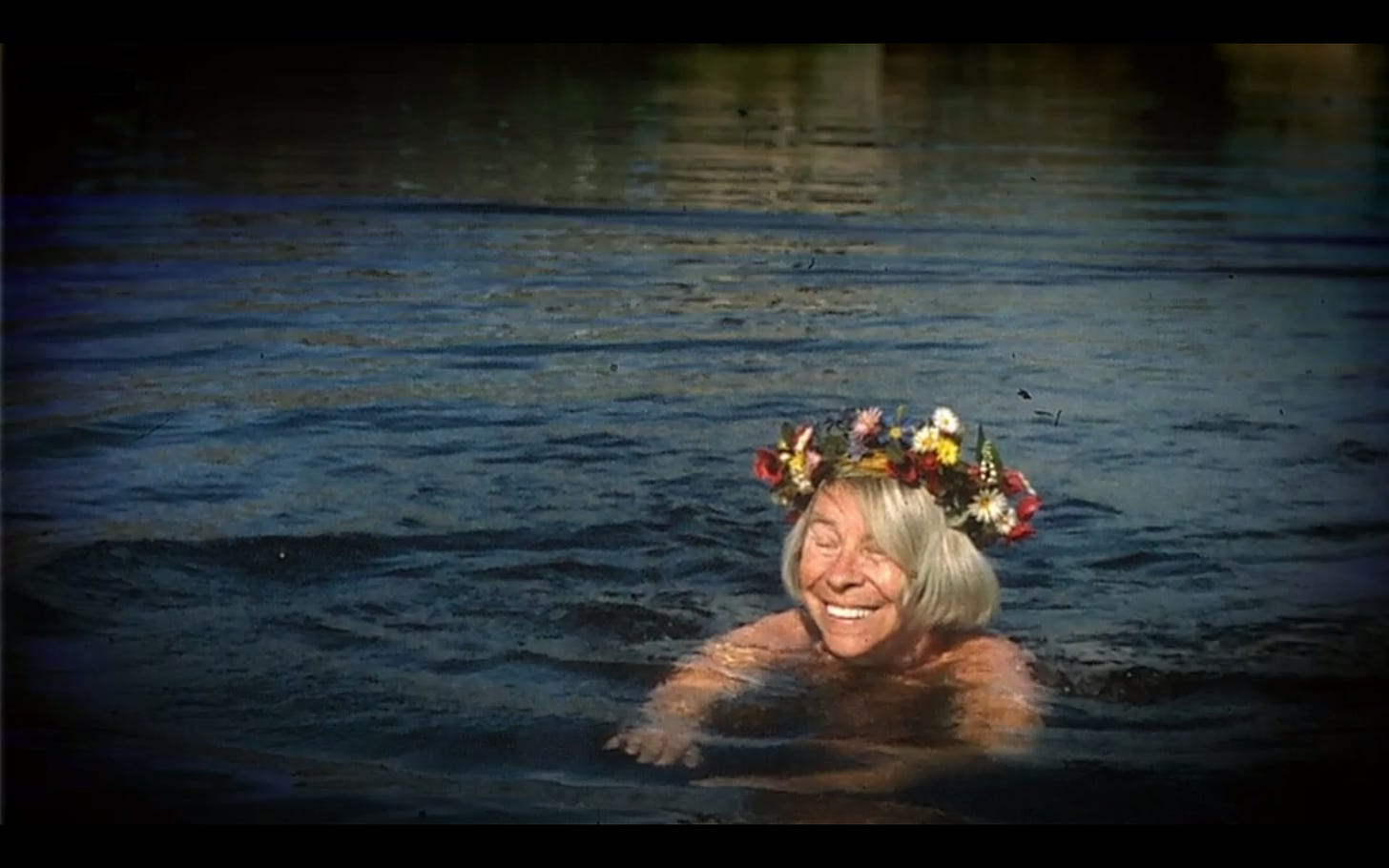 An older white woman with straight grey hair in a bob style with a crown of flowers, swims naked with a big smile on her face in a dark lake.