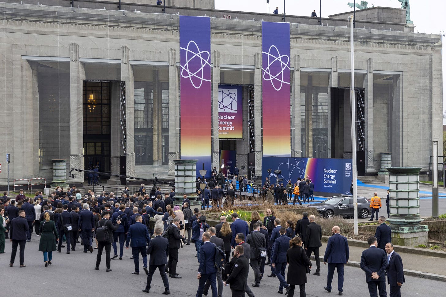 A crowd of people wearing business suits walks towards a convention center building decorated with banners for the nuclear energy summit.