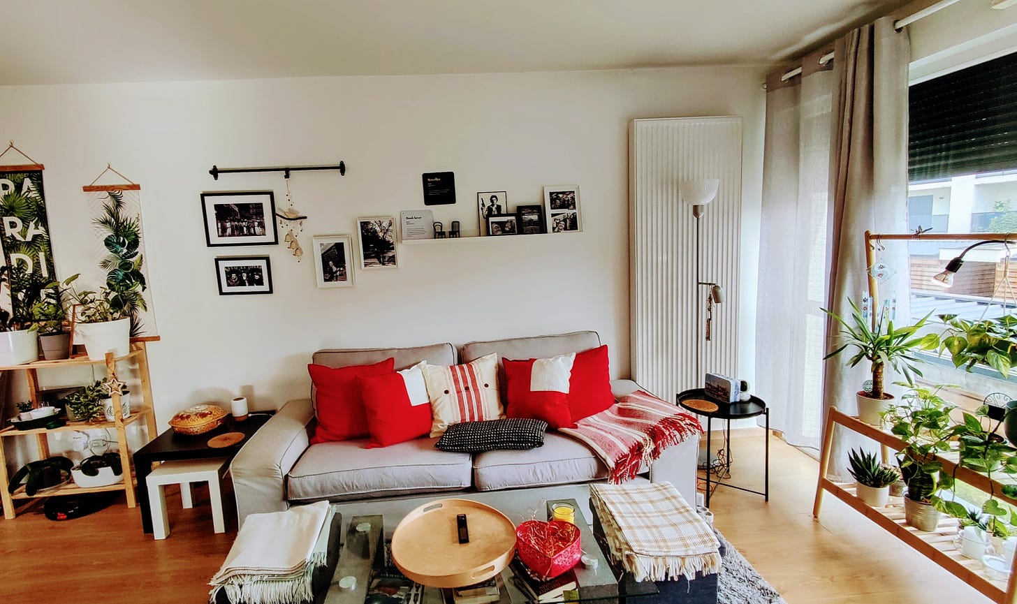 Picture of my living room. There’s a three-seat light gray couch, red and white throw pillows and a red and white striped blanket. In the wall behind the couch there’s a small gallery of black and white photos, and there are several plants in wooden shelves both to the right and the left of the room.