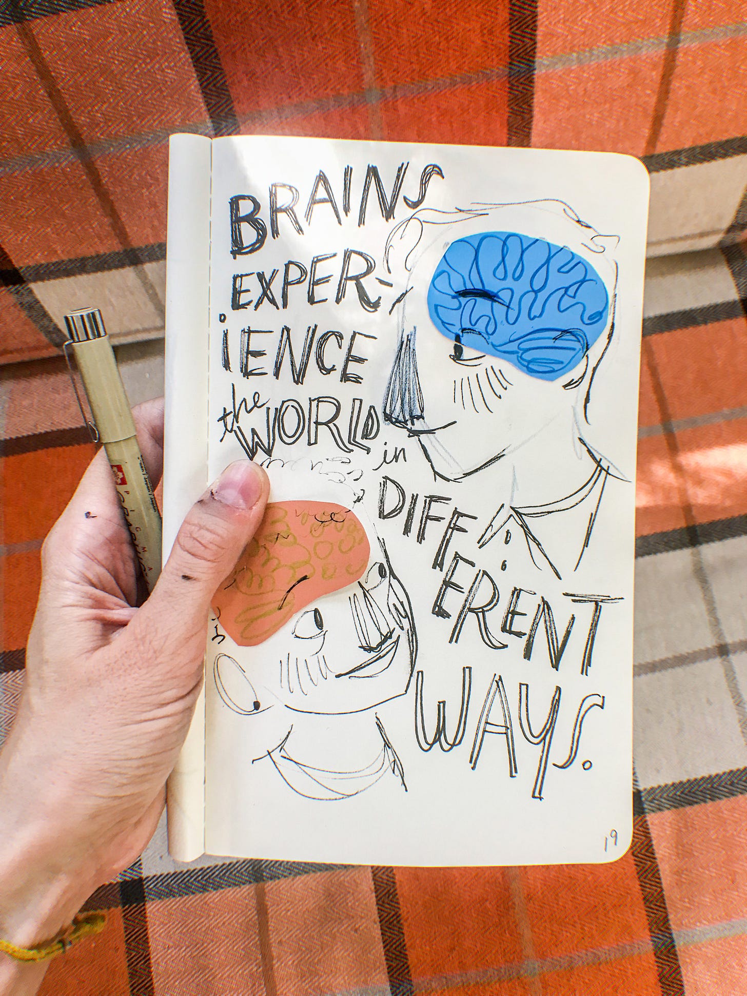 White ink stained hand holds sketchbook. Hand written text reads brains experience the world differently.
