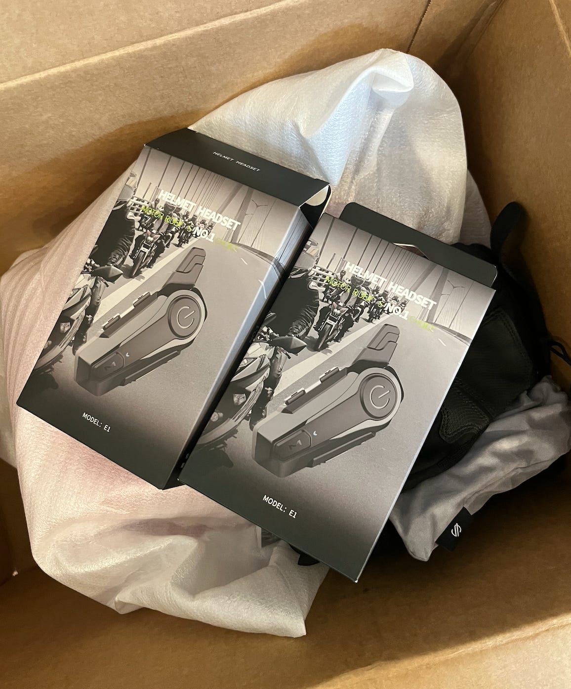 A look inside an open shipping box. Two packages for bluetooth "Helmet Headsets" rest on top of white protective cover for Amber's motorcycle helmet.
