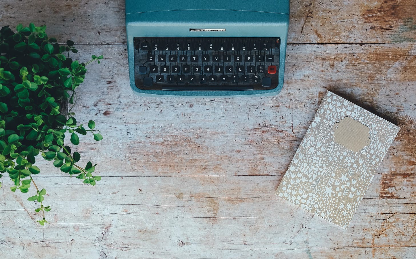 teal typewriter on a wooden desk with a potted plant on the left and a journal on the right