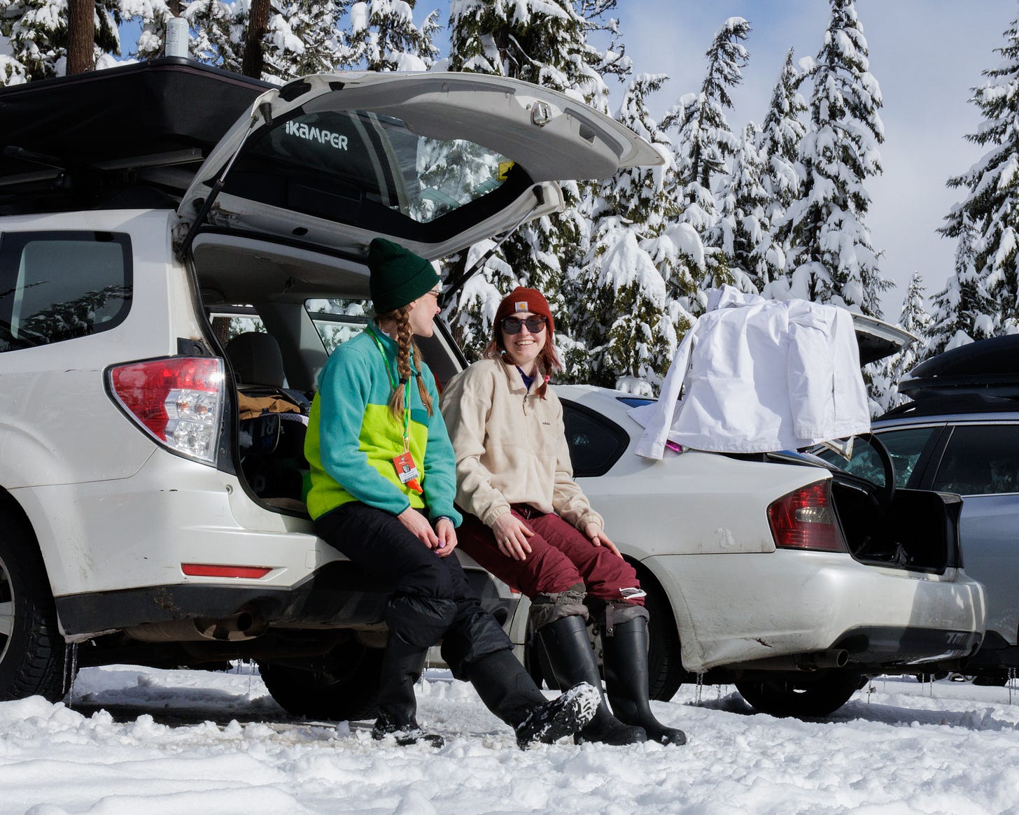 Two people sitting in the open trunk of a sedan at a ski area, talking.