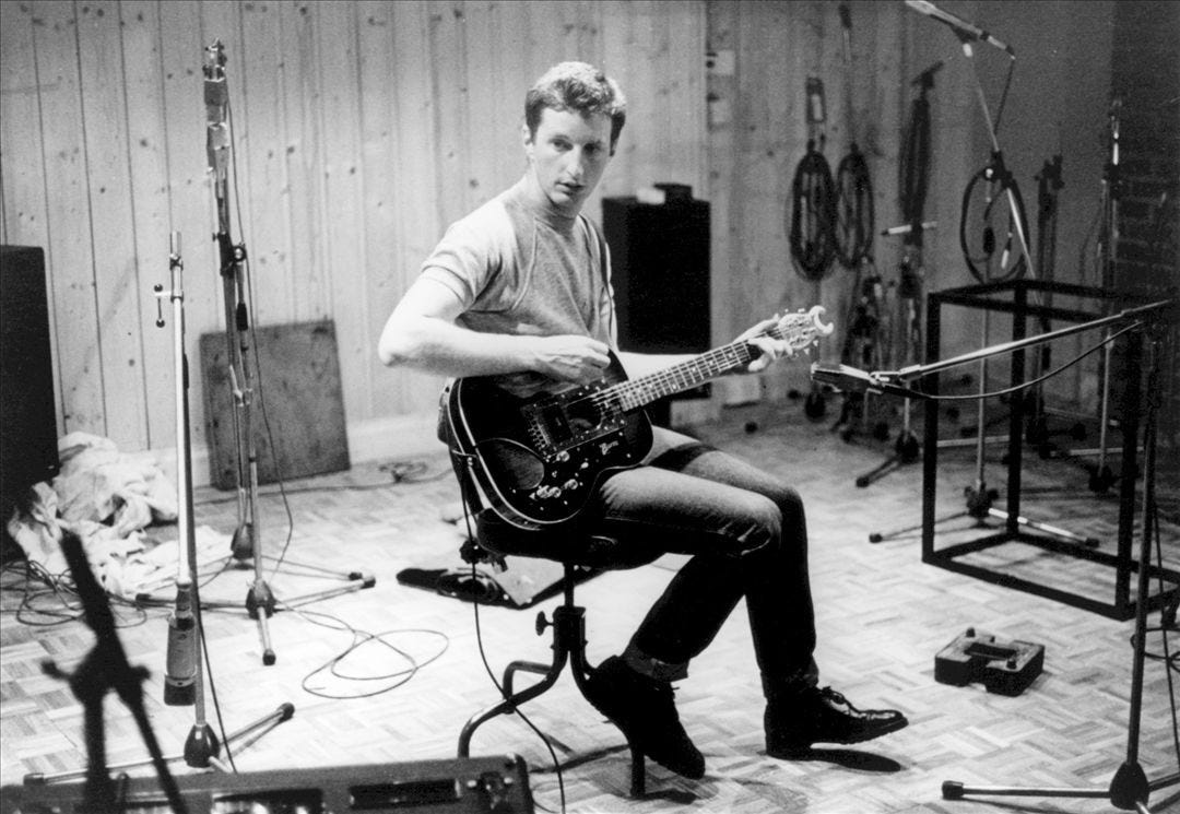 black and white photo of a white man (billy bragg) sitting on a stool with a guitar perched on one leg, preparing to start a song