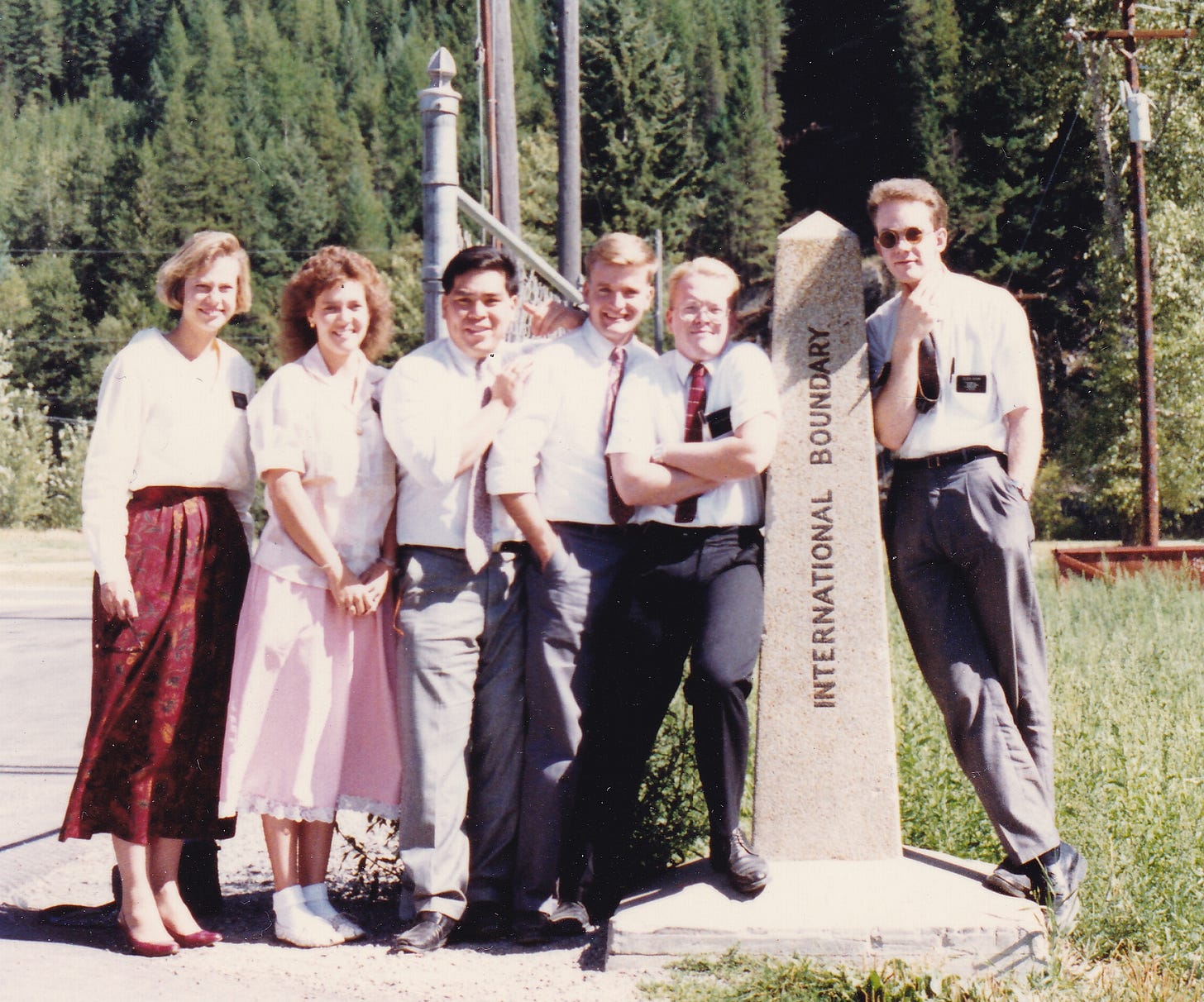 Five Mormon missionaries (two female, three male) crowd against the left side of a stone pylon marked "INTERNATIONAL BOUNDARY," while one lone male missionary leans against the right side. Behind them a wooded hillside rises with a corridor clearcut through the trees.