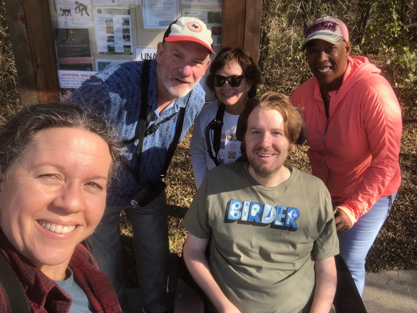 Celeste takes a selfie with four other birders in front of an information kiosk for a woodsy neighborhood trail. They smile in the sunlight while wearing a mix of binoculars, ball caps, and sunglasses. The word “Birder” appears on Eric’s T-shirt; the letters are formed by assistive equipment like prosthetic legs and hearing aids.