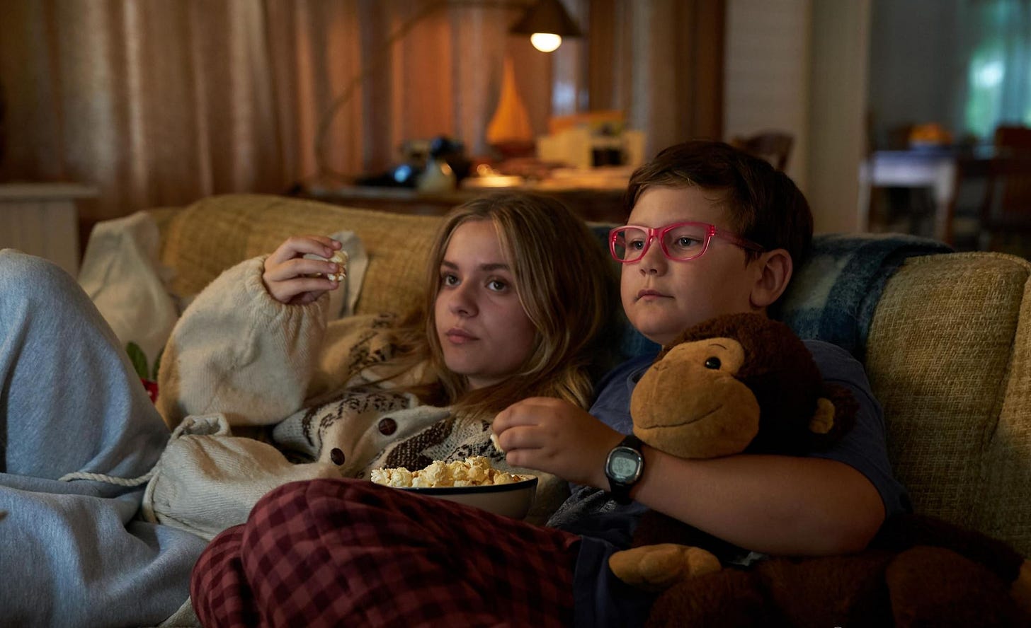 Elliott and her younger brother eat popcorn on the couch while watching TV.