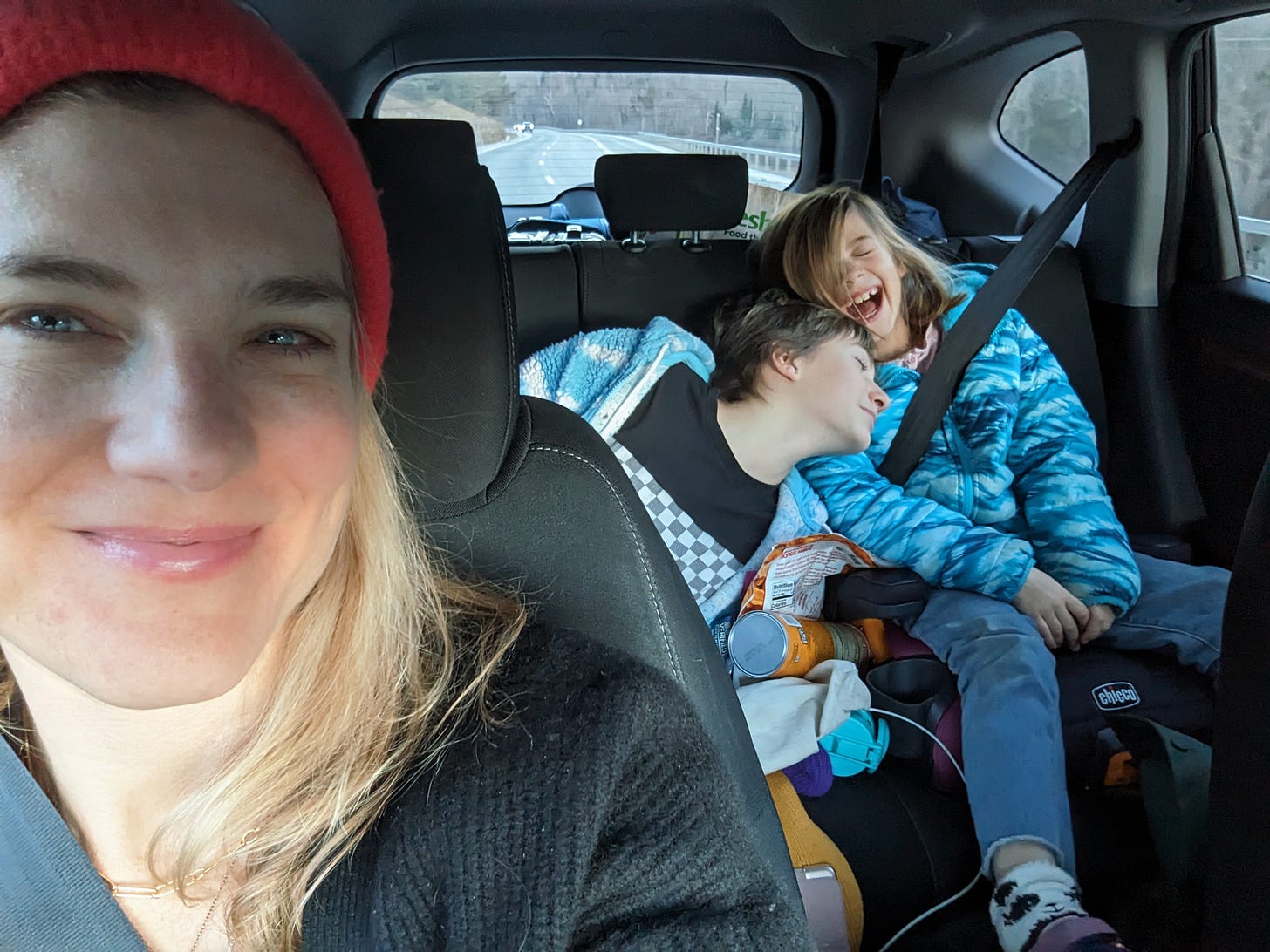 A selfie blonde woman looking at the camera at two children in the backseat of a car