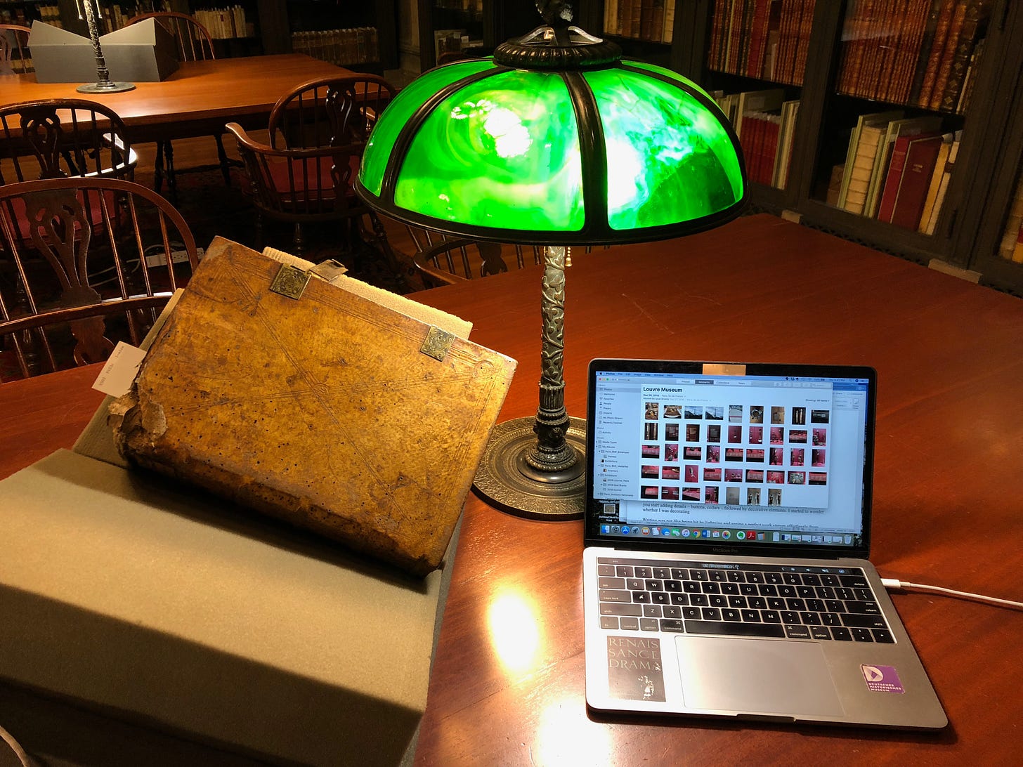 An old book bound with clasps sits on cushions on a wooden library table lit by a decorative desk lamp next to a Mac Powerbook.