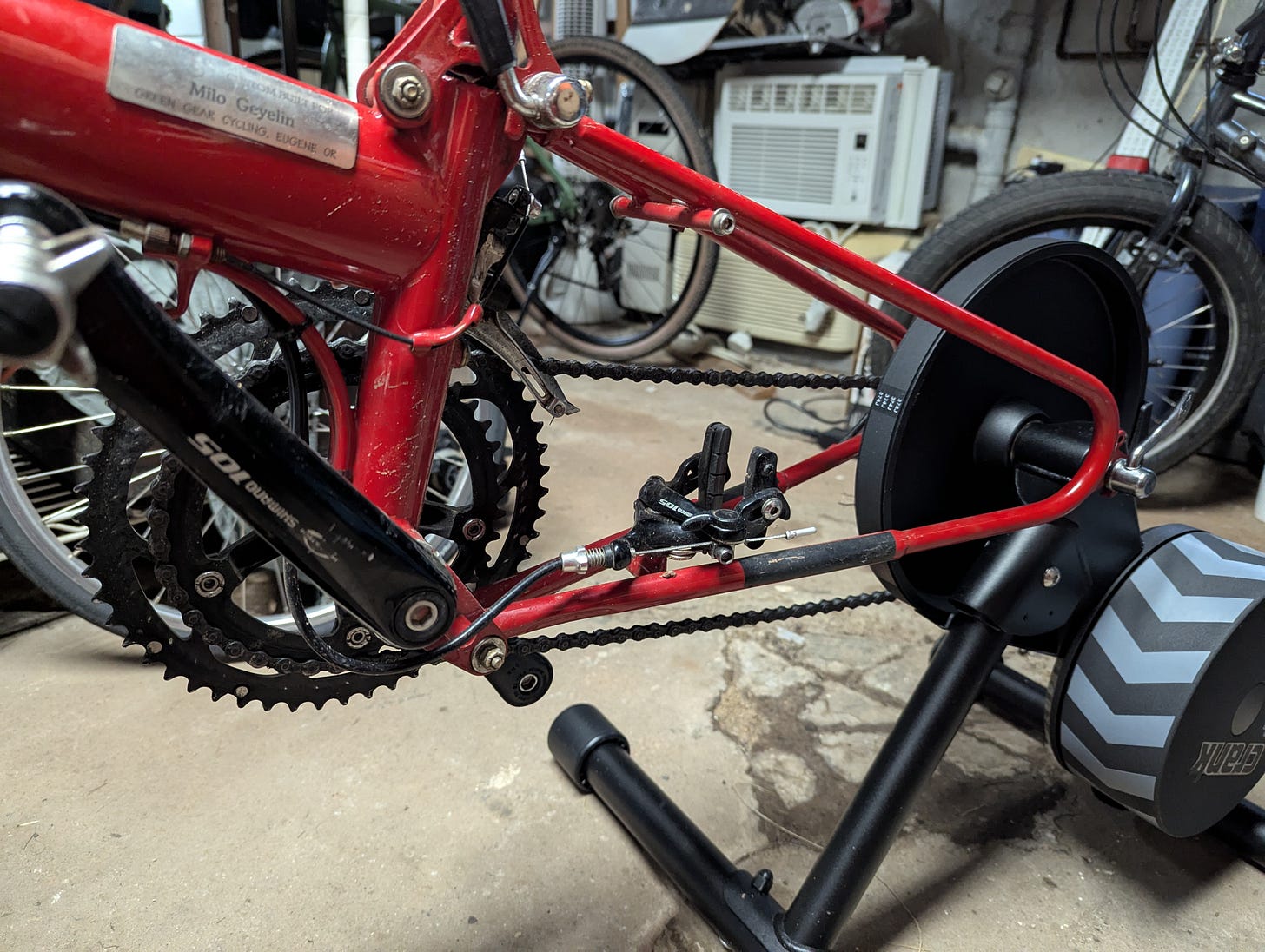 Close up photo of a bicycle on a stationary trainer. The rear wheel of the bicycle is removed and in it's place is the trainer. The trainer has a stable, tubular base, a heavy, round flywheel, and another round wheel that sits where the bicycle's rear tire typically sits.