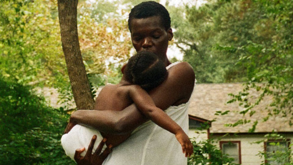 A Black woman in white holds a sleeping baby in front of a house and trees.