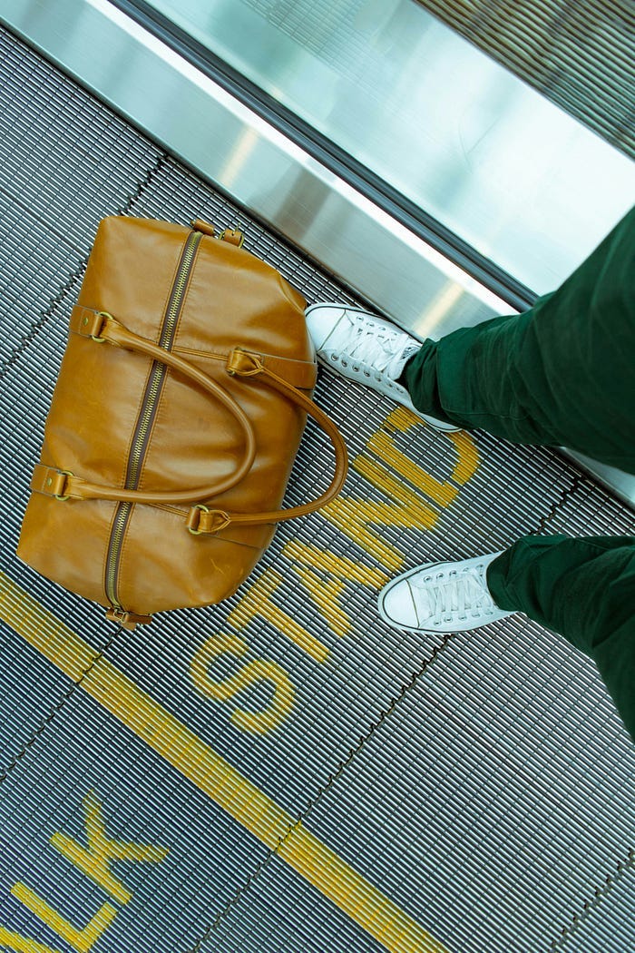 Photo of brown travel case near a person wearing black trousers and white shoes.