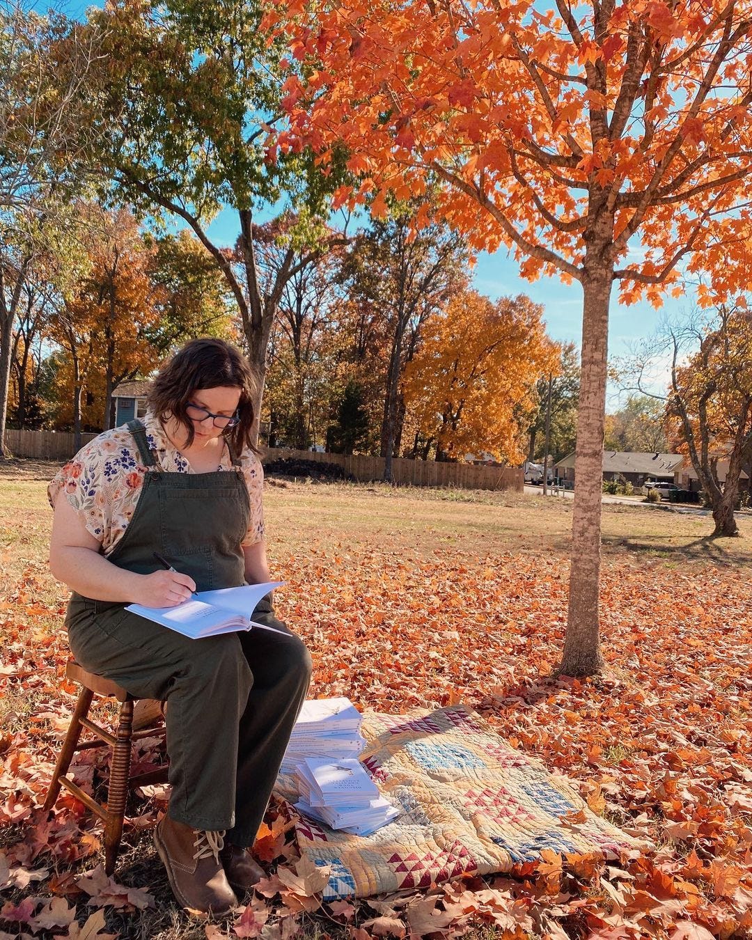 I'm sitting under an orange maple tree wearing olive overalls and signing a stack of books. I have pale skin, short brown. hair, and green glasses.