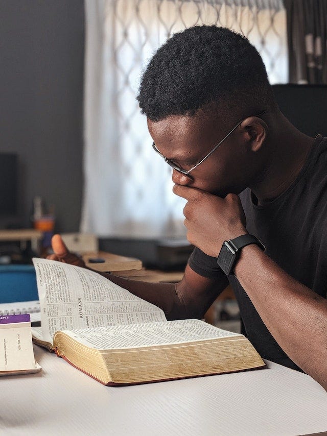 Black man with close-cropped fade in black t-shirt with black watch on reads a Bible open to the book of Romans.