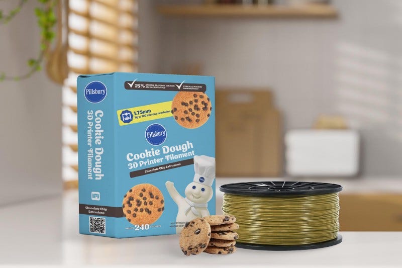 A large square box labelled Pillsbury Cookie Dough 3D Printer Filament on a kitchen counter next to a spool of cookie coloured filamant and a stack of chocolate chip cookies.