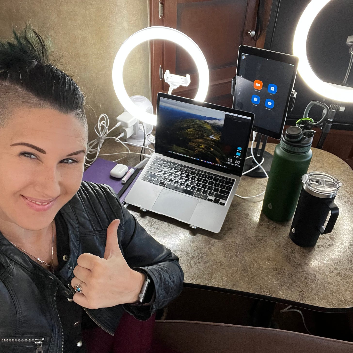  Lyric Rivera, giving a thumbs up. They are wearing a black leather jacket and black top, sitting in their RV, as they sit behind their laptop, iPad, and other devices for broadcasting live to the world. 