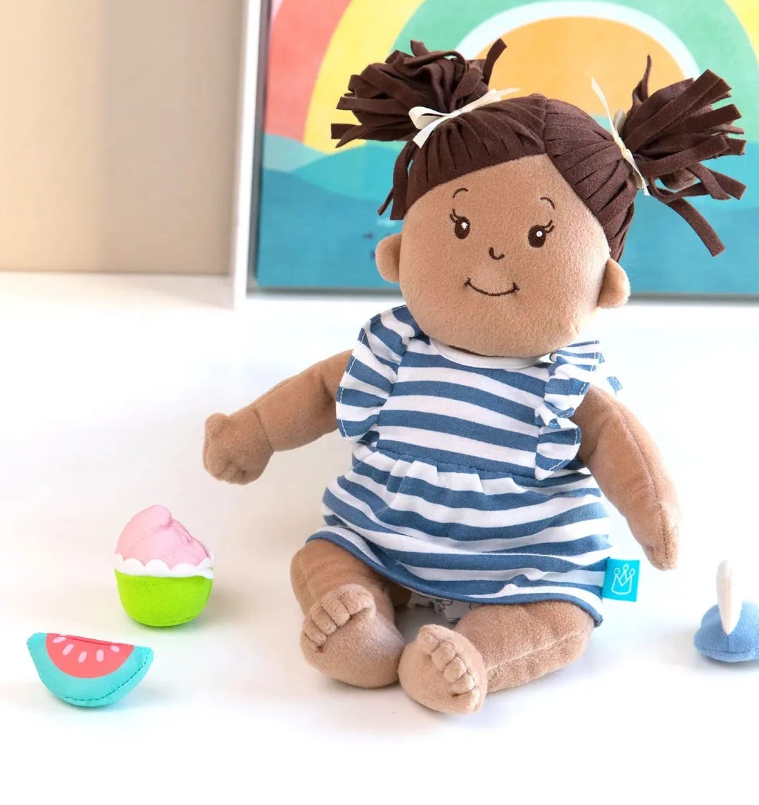 A fabric doll with tan skin and dark brown hair sits in a white and blue striped dress with toy accessories strewn around her.