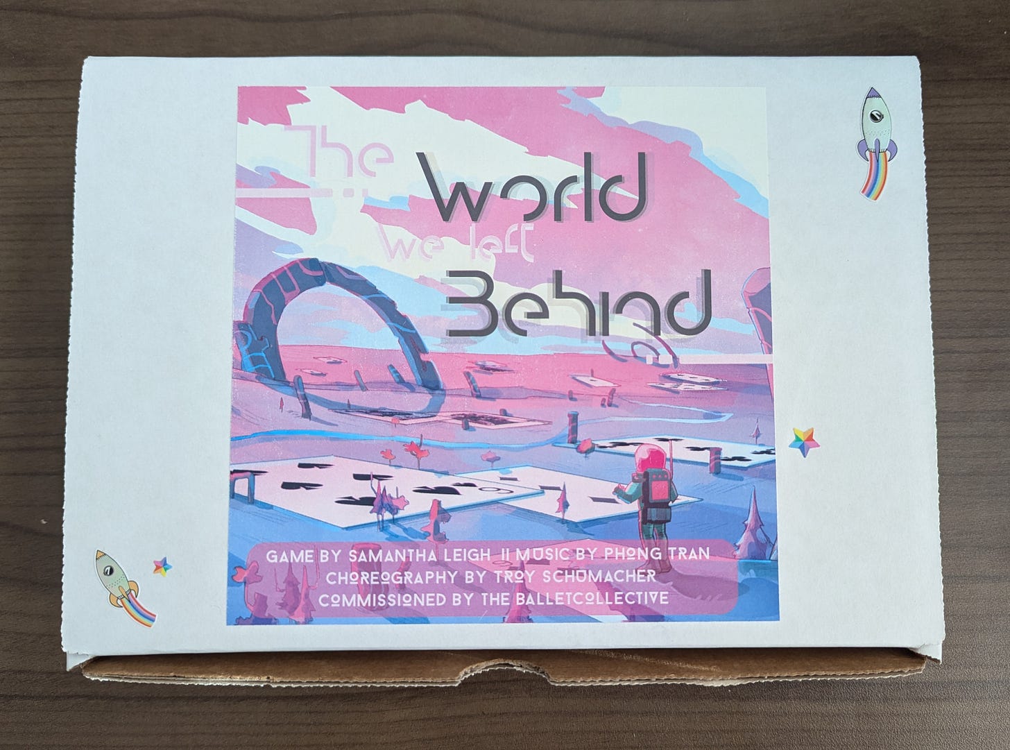 The box for The World We Left Behind bundle. A white cardboard box with a large sticker of the cover art of TWWLB. A couple smaller stickers of spaceships and starts dot the box.