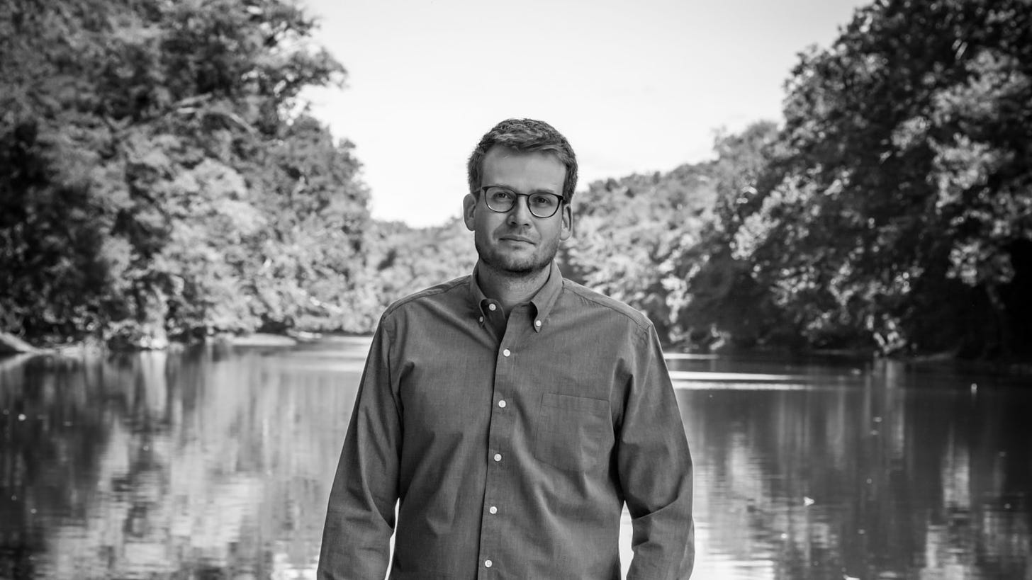 Author and Awesome Coffee Club co-founder John Green standing in front of a lake looking at the camera. Credit Marina Waters, 2021