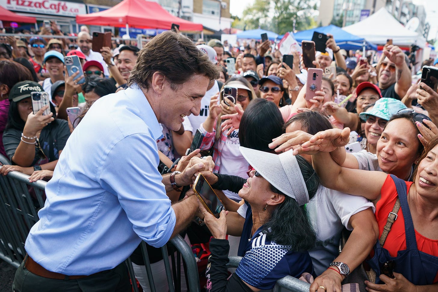 Prime Minister Justin Trudeau is shaking hands with a person. A large crowd of people is standing in front of him and beside him.
