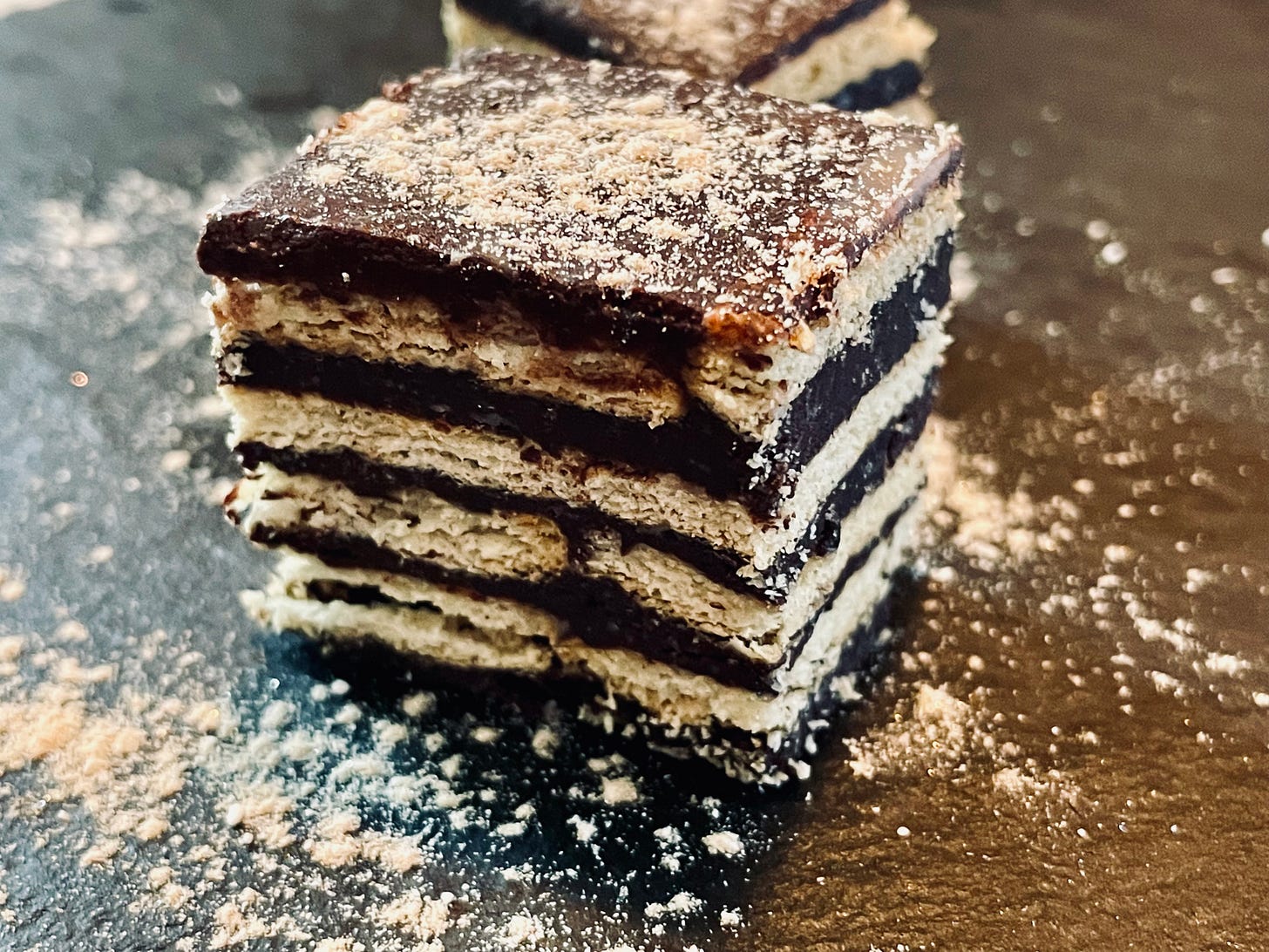 Close-up image of a single Kalter Hund Praliné cube, showcasing alternating layers of chocolate and biscuits, topped with a light dusting of powdered sugar. The dessert's smooth texture and defined layers create a visually appealing, elegant look. Close-up image of a single Kalter Hund Praliné cube, showcasing alternating layers of chocolate and biscuits, topped with a light dusting of powdered sugar. The dessert's smooth texture and defined layers create a visually appealing, elegant look.
