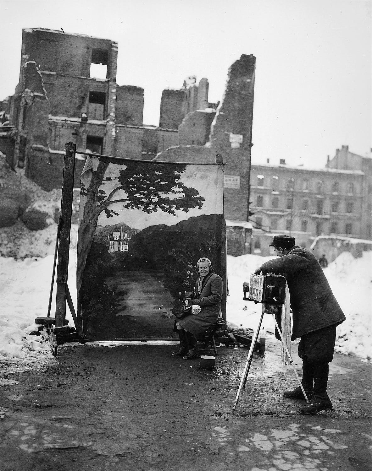 A man is taking a photograph of a seated woman. She is sitting in front of a painted backdrop featuring an idyllic rural scene. The real backdrop behind that is a bombed out cityscape.