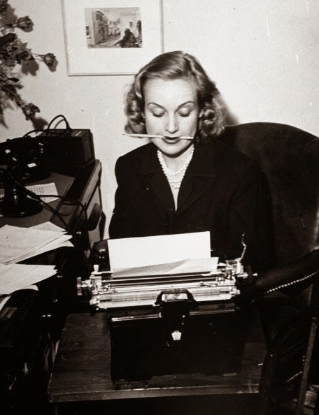 Black and white photo of a woman sitting at a desk in front of a typewriter, a pencil in her mouth