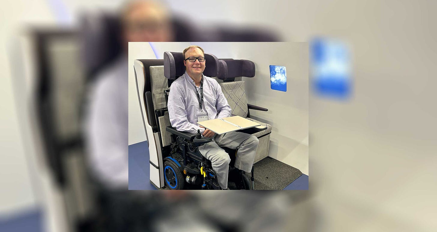 John seated in a wheelchair in a wheelchair securement space for airplanes, with the tray table extended over his lap.