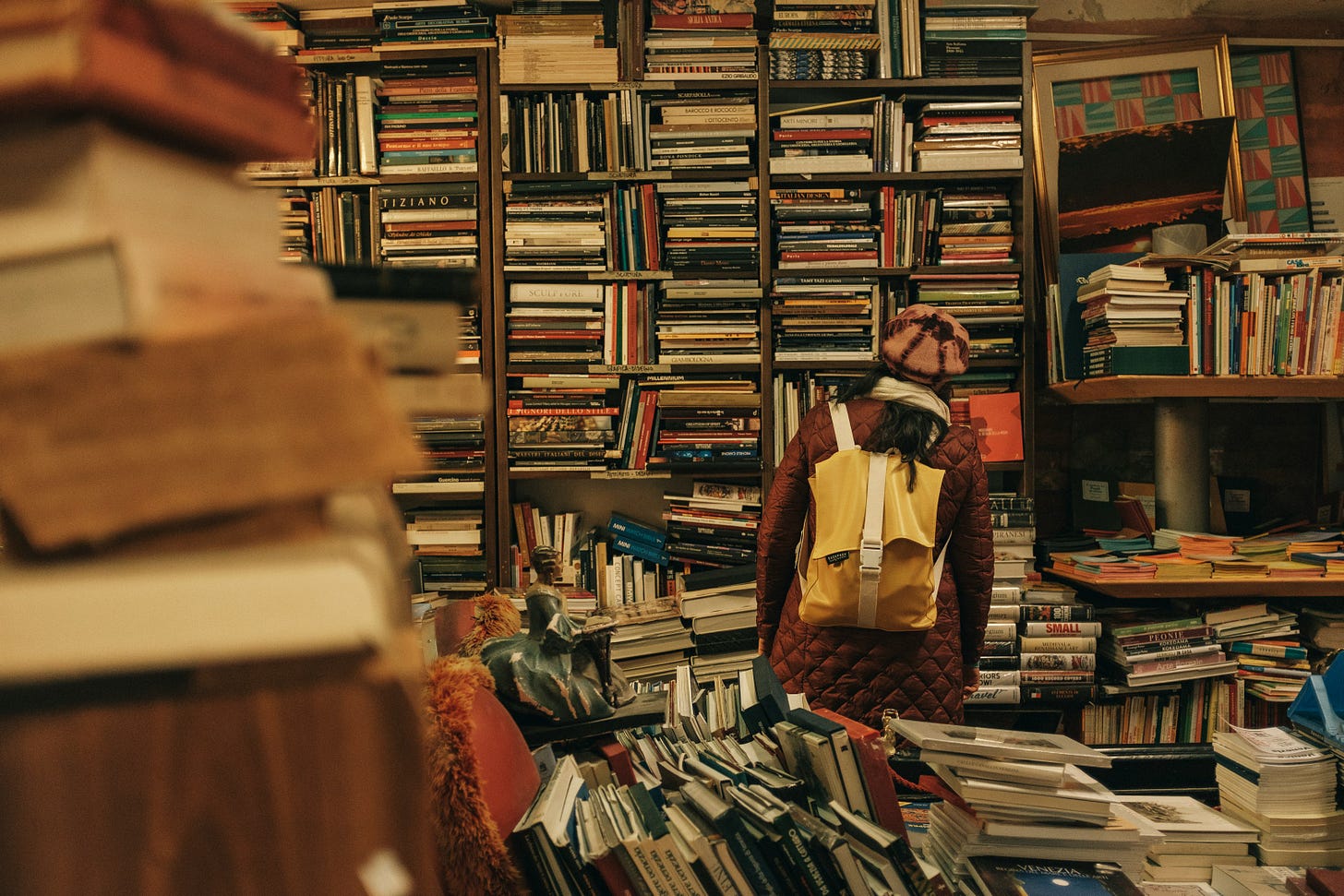 A photo of a second hand book store, shelved piled high with old books are everywhere and a lady with a yellow backpack is looking intently at one of the shelves, with her back to the camera.
