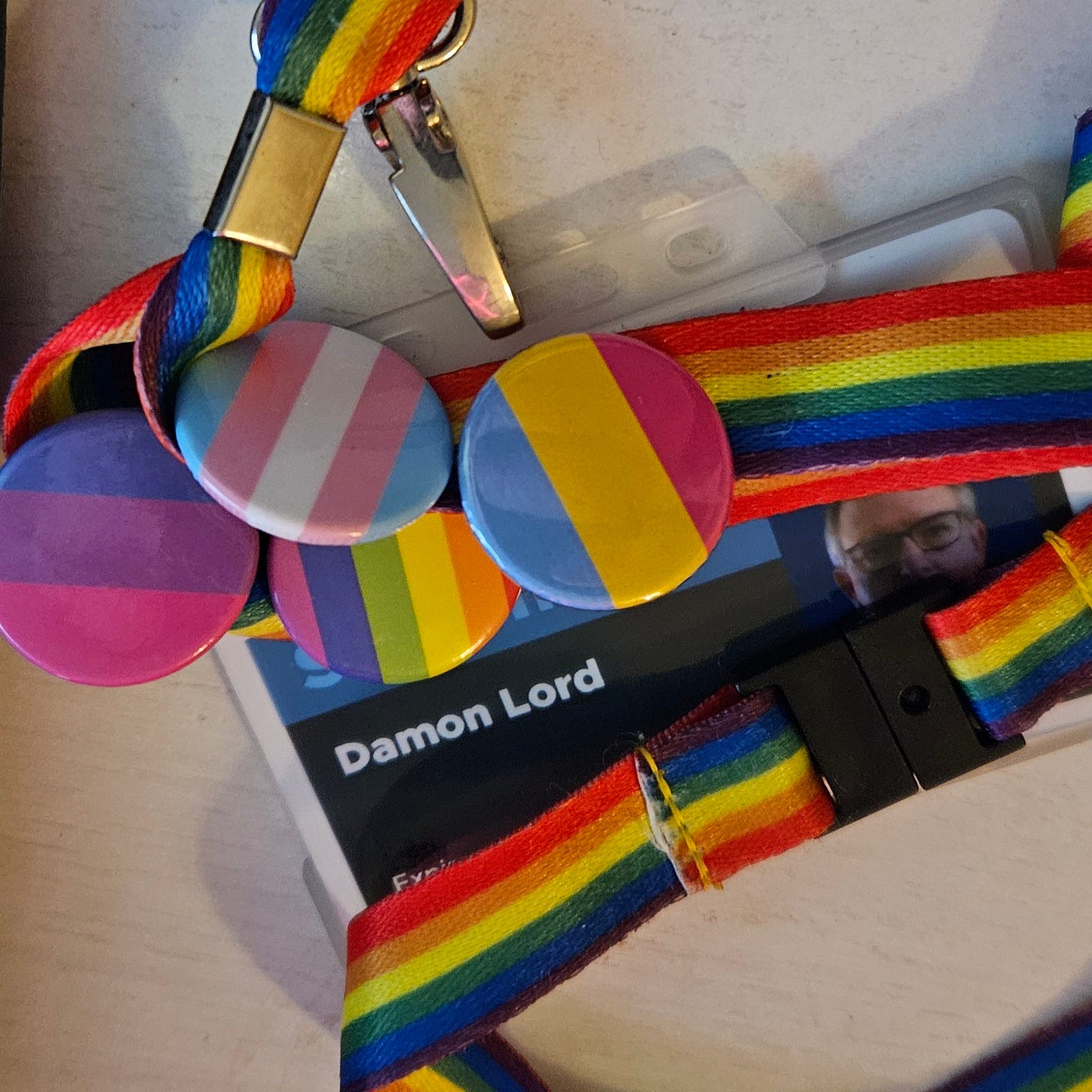 Damon Lord's ID card, with a rainbow lanyard draped across it, mostly obscuring the card except for the name Damon Lord. Attached the the lanyard are four pin badges displaying the trans flag, the rainbow flag, the bisexual flag, and the pansexual flag. It lies upon a white table surface.