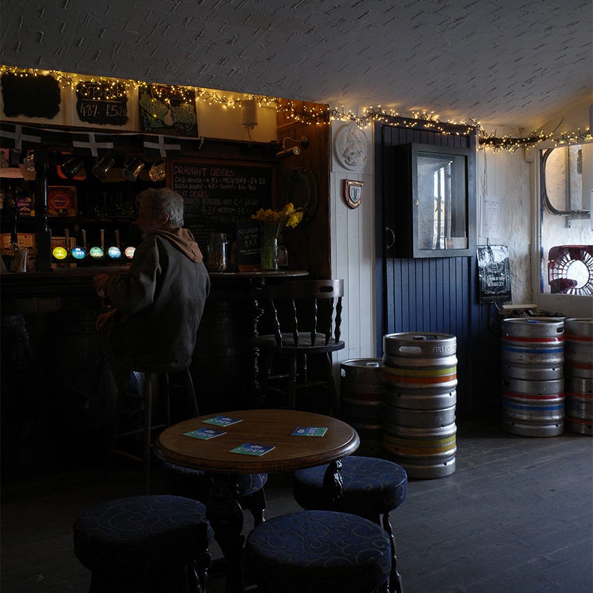 The interior of a Cornish pub with low light, stacked beer casks, fairy lights, and a shadowy figure sitting at the bar.