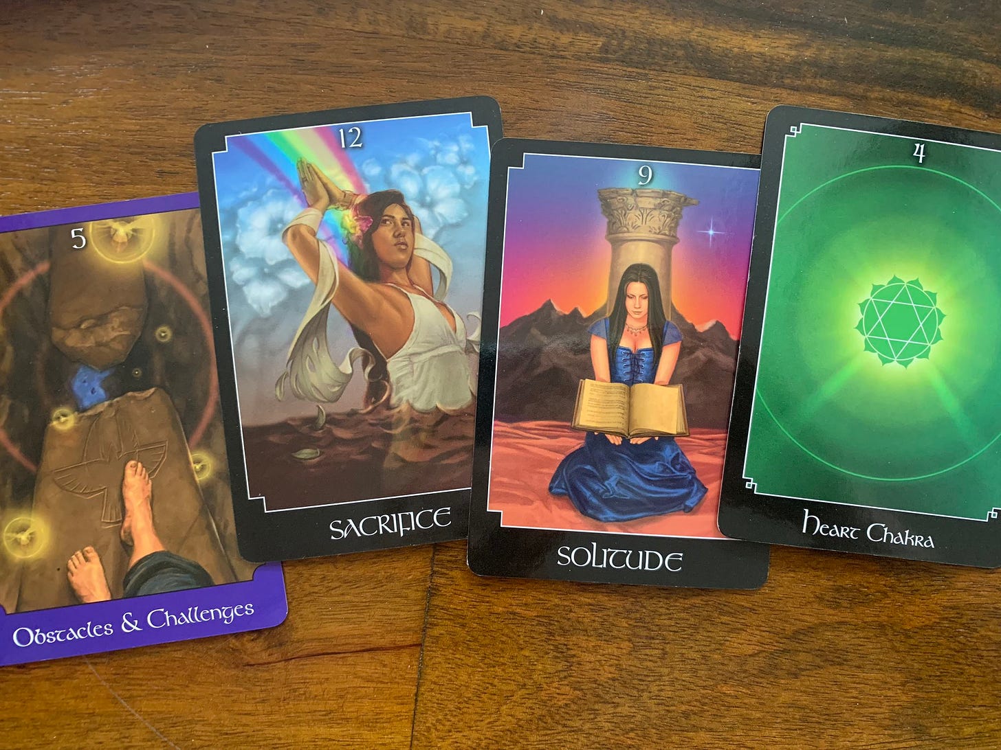 Four tarot cards on a wooden table. "Obstacles and Challenges." "Sacrifice." "Solitude." And "Heart Chakra."