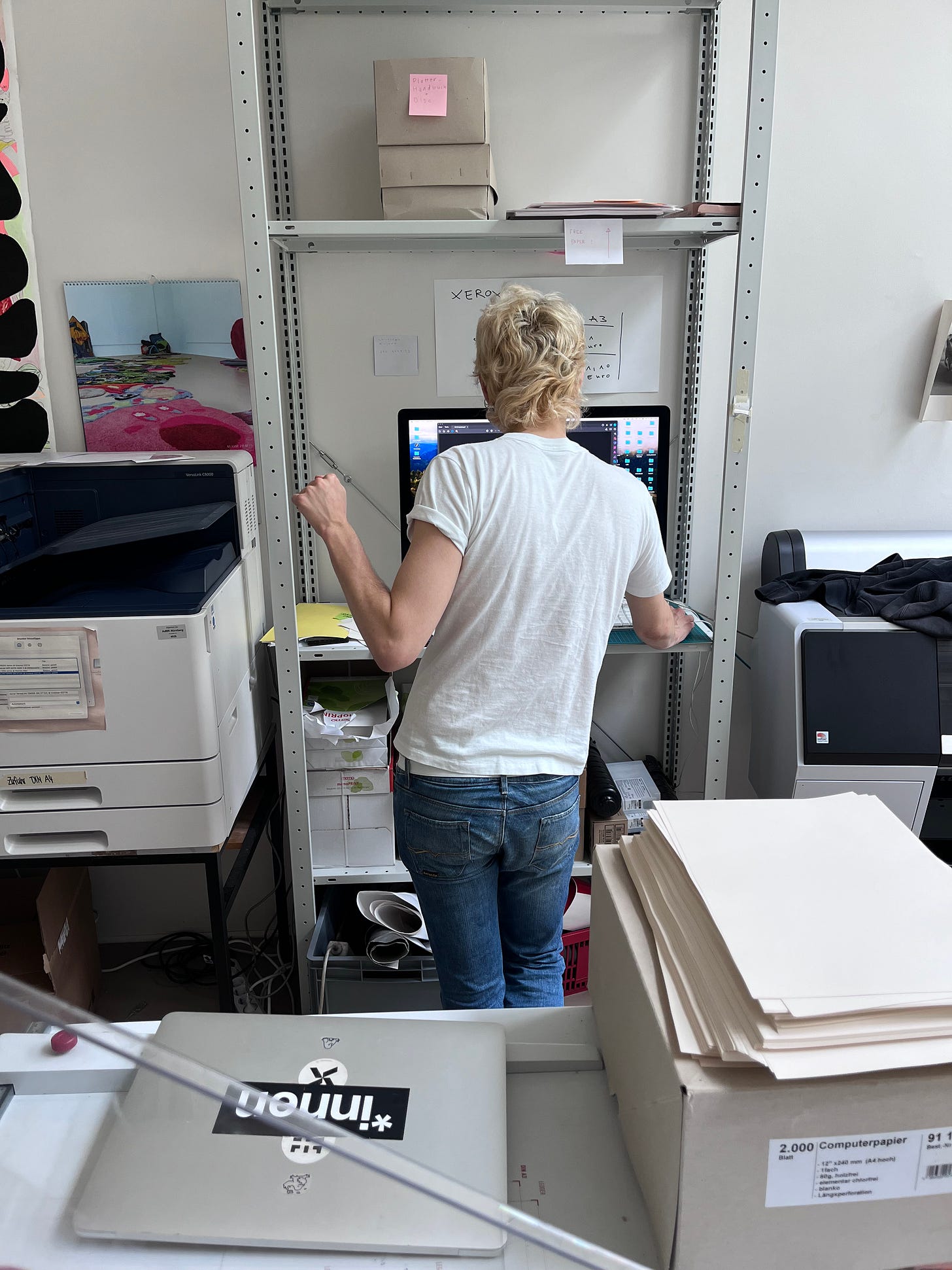 Mark is pictured here from behind, holding up a fist with his left hand, dealing with a desktop computer with his right hand. Mark has a queer looking blonde mullet and is wearing a white t shirt and bell bottom jeans. The printer is to the left of mark, and other tools and pages and shelves of the graphic design department are scattered around the image. Mark's laptop is on the table at the bottom of the image, and it has a sticker on it with the words *innen.