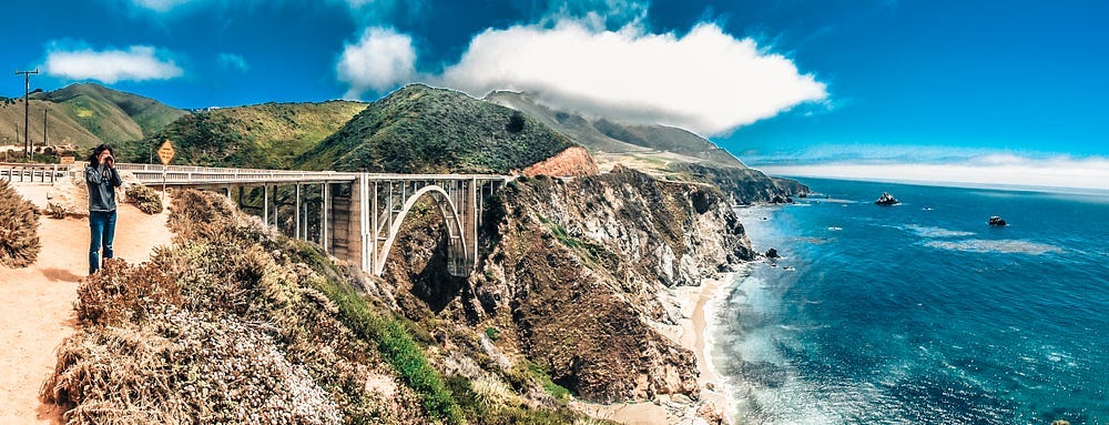 Man looks out over Pacific Ocean with dramatic Bixby Bridge behind.