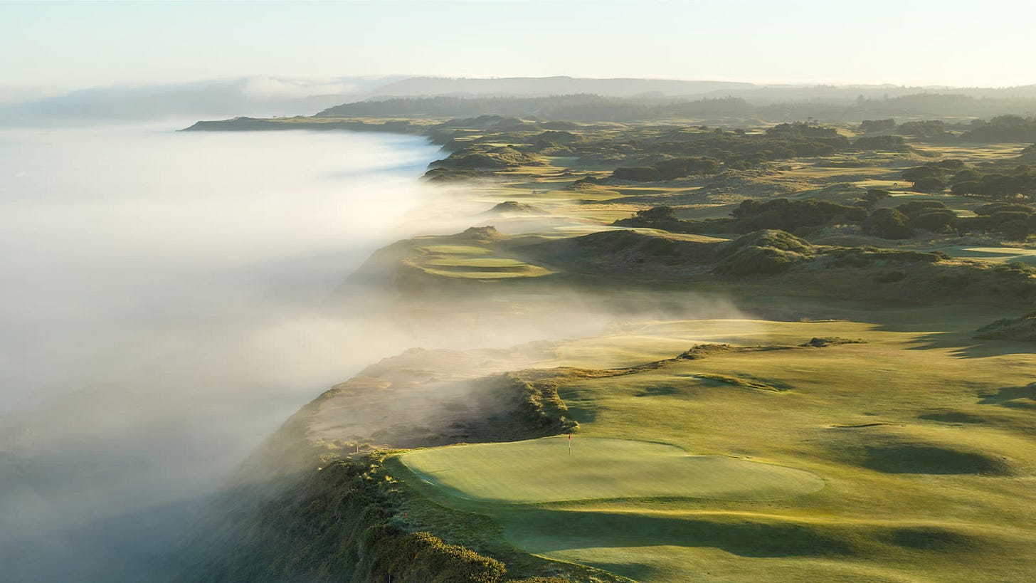 Aerial view of Bandon Dunes Golf Resort during a misty morning, showing emerald green fairways winding through natural dunes. Rolling fog dramatically spills over the coastal cliffs, creating a mystical atmosphere where the Pacific Ocean meets the rugged Oregon coastline. Aerial view of Bandon Dunes Golf Resort during a misty morning, showing emerald green fairways winding through natural dunes. Rolling fog dramatically spills over the coastal cliffs, creating a mystical atmosphere where the Pacific Ocean meets the rugged Oregon coastline.