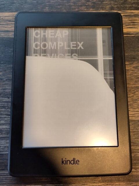 Photo of a Kindle book-reading tablet. It  shows the cover of my novella Cheap Complex Devices, but the image is all messed up, as if the screen is cracked and the machine is broken. 