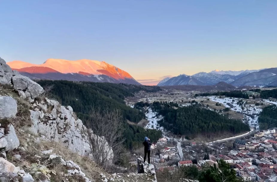 A person stands on a rocky hill overlooking a valley and snow-capped mountains, capturing the beauty of nature during sunrise.