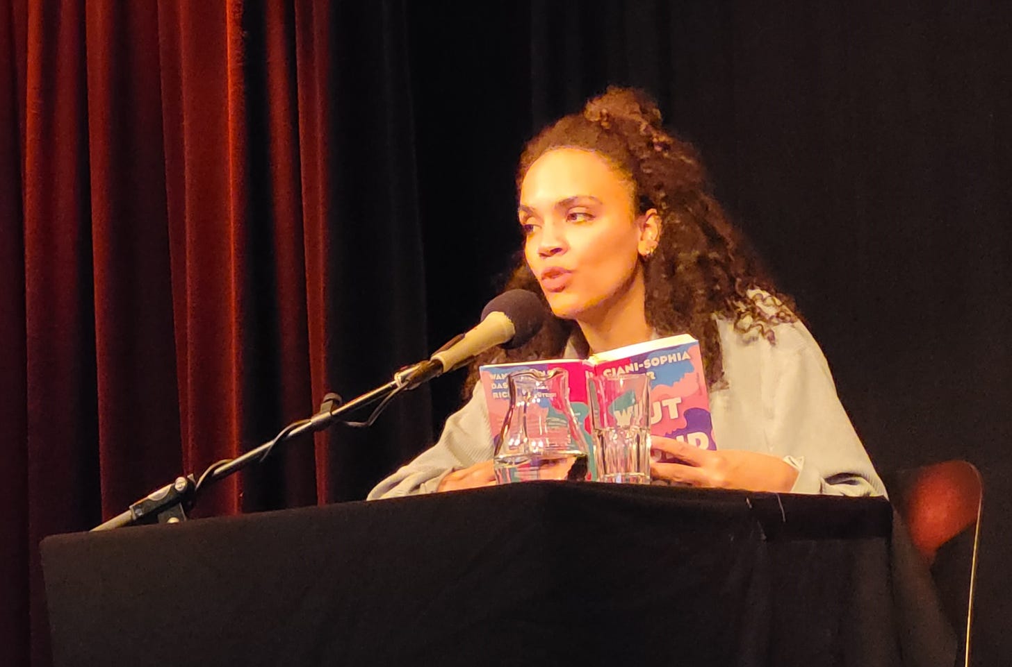 Ciani-Sophia Hoeder sitzt vor dunklem Hintergrund an einem schwarzen Tisch mit Mikrofon. In Händen hält sie ihr Buch „Wut und Böse“.
