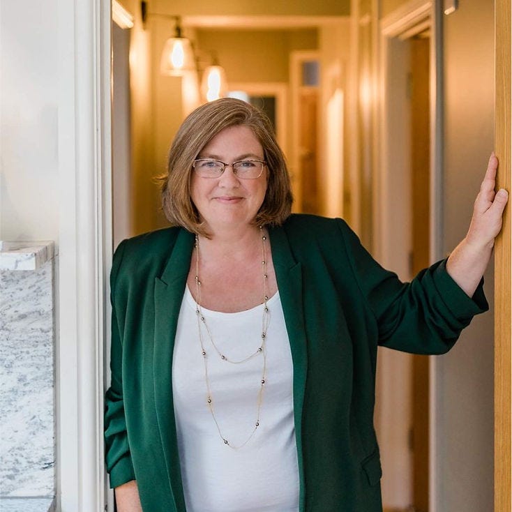 Lorna helps in a white top and green jacket, standing in a doorway