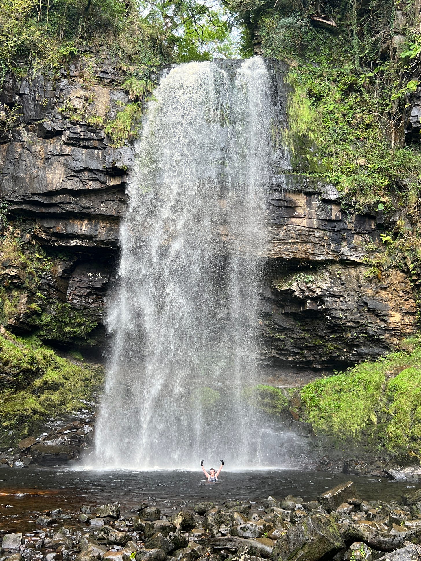 A very very tall waterfall cascades down into a pool where a very small but very happy swimmer waves at the camera.