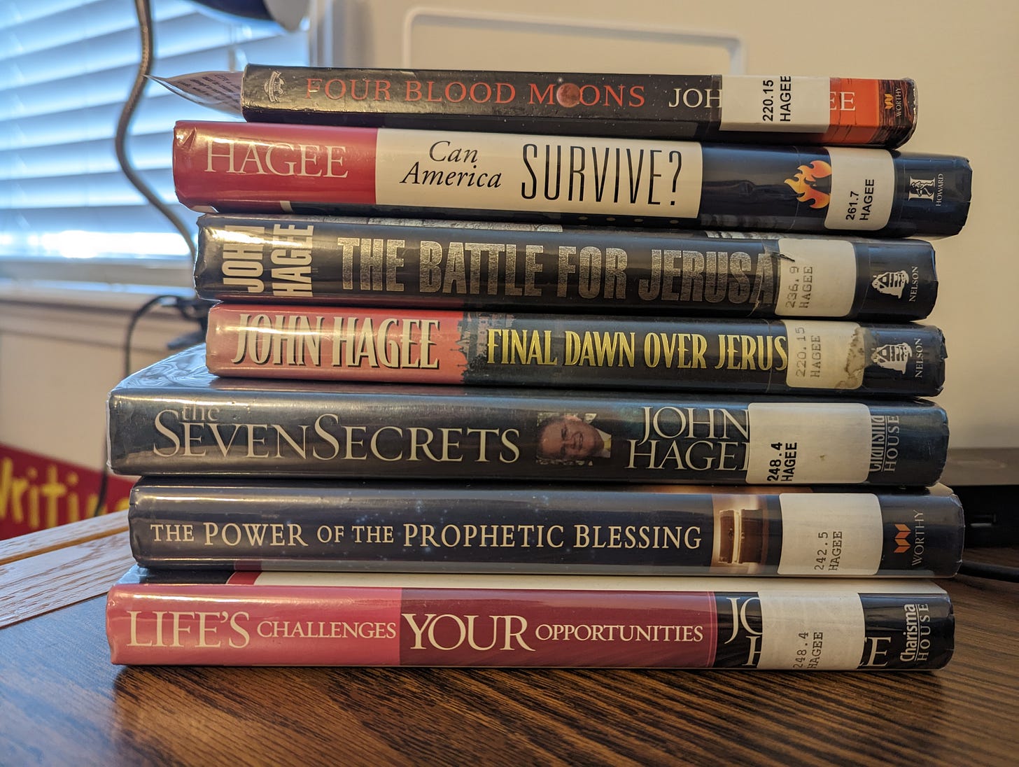 A stack of Hagee's books on a desk: Four Blood Moons, Can America Survive, The Battle for Jerusalem, Final Dawn Over Jerusalem, The Seven Secrets, The Power of the Prophetic Blessing, and Life's Challenges Your Opportunities.