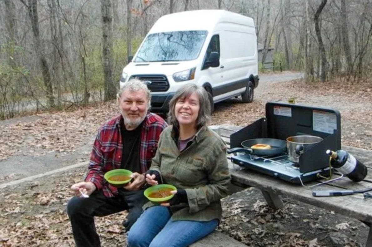 A man and a woman eat on a bench in the woods, a white van parked in the background