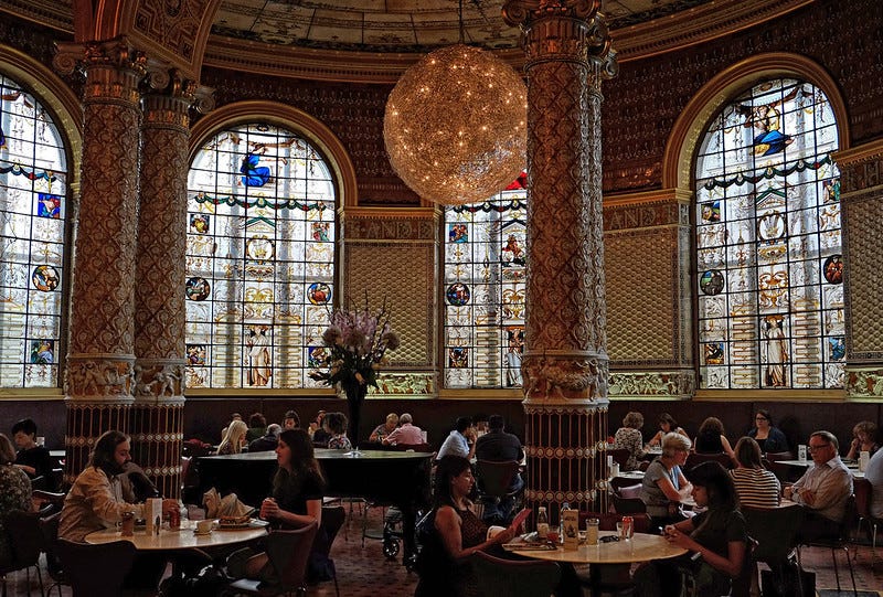 People drink tea and chat at tables in a grand museum cafe, with ornate carved pillars and a spherical pendant lamp.