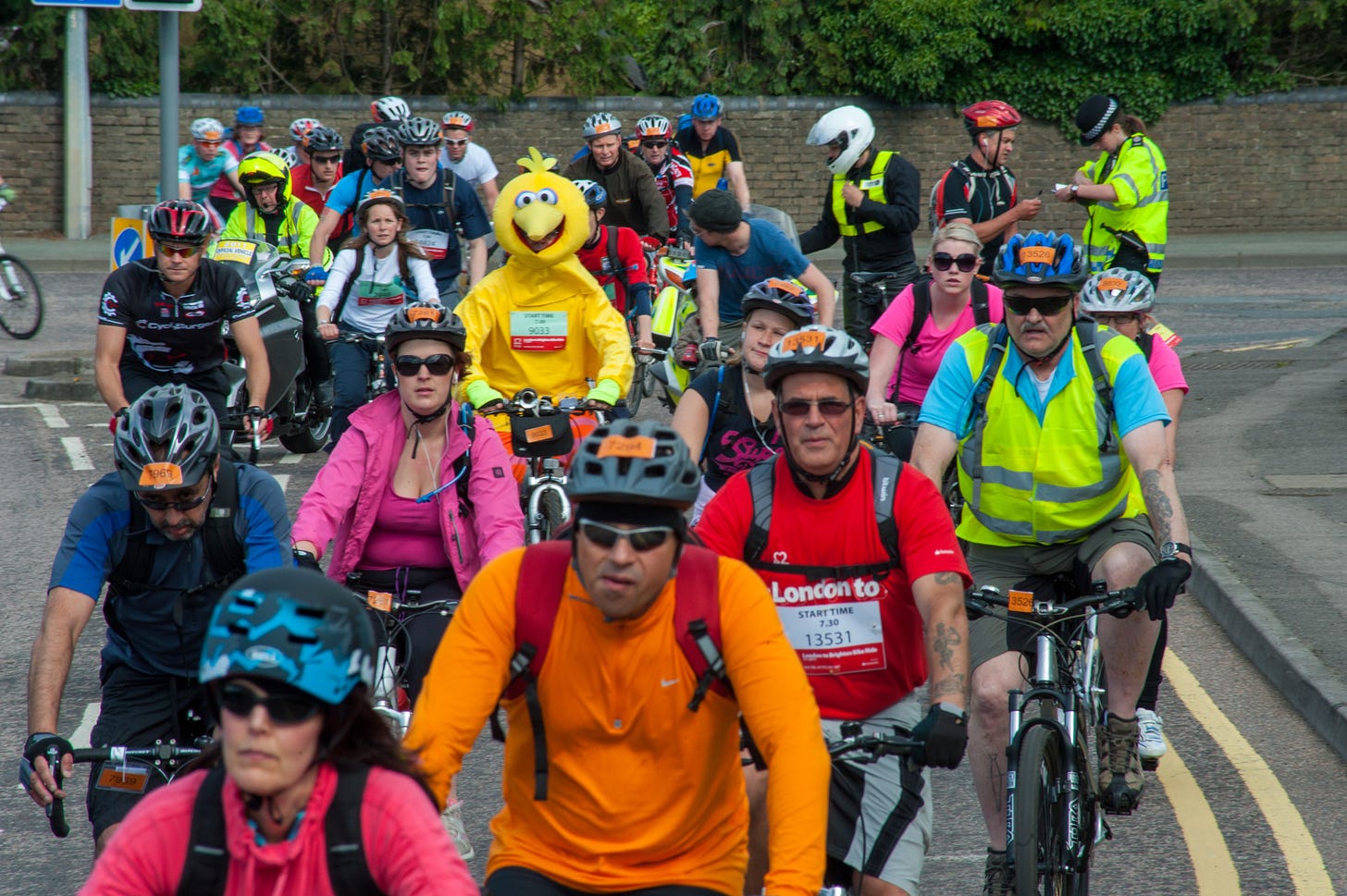 A colorful group of cyclists are driving torwards the camera. While most people are wearing standard cycling attire, one person is masked as Big Bird of Sesame Street.