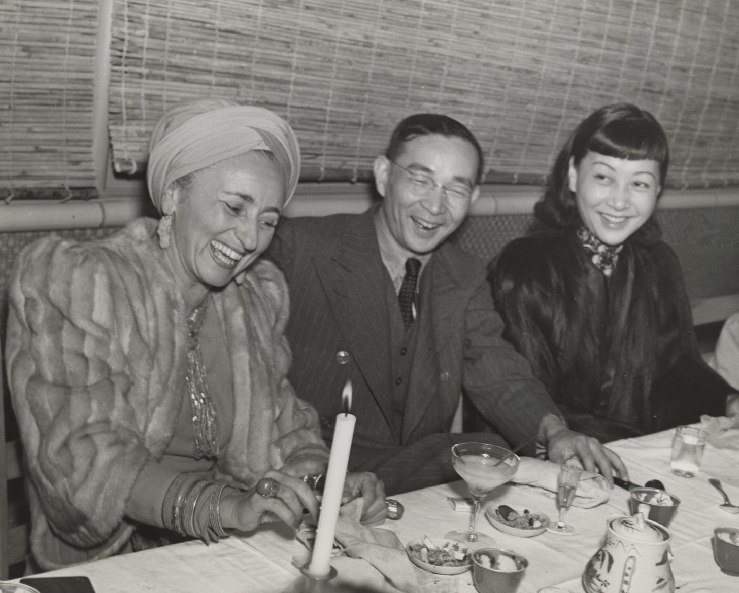 Bernardine Fritz in furs and turban and jewels, Lin Yutang in between her and AMW also wearing a dark fur coat, sitting at a dinner table and laughing together