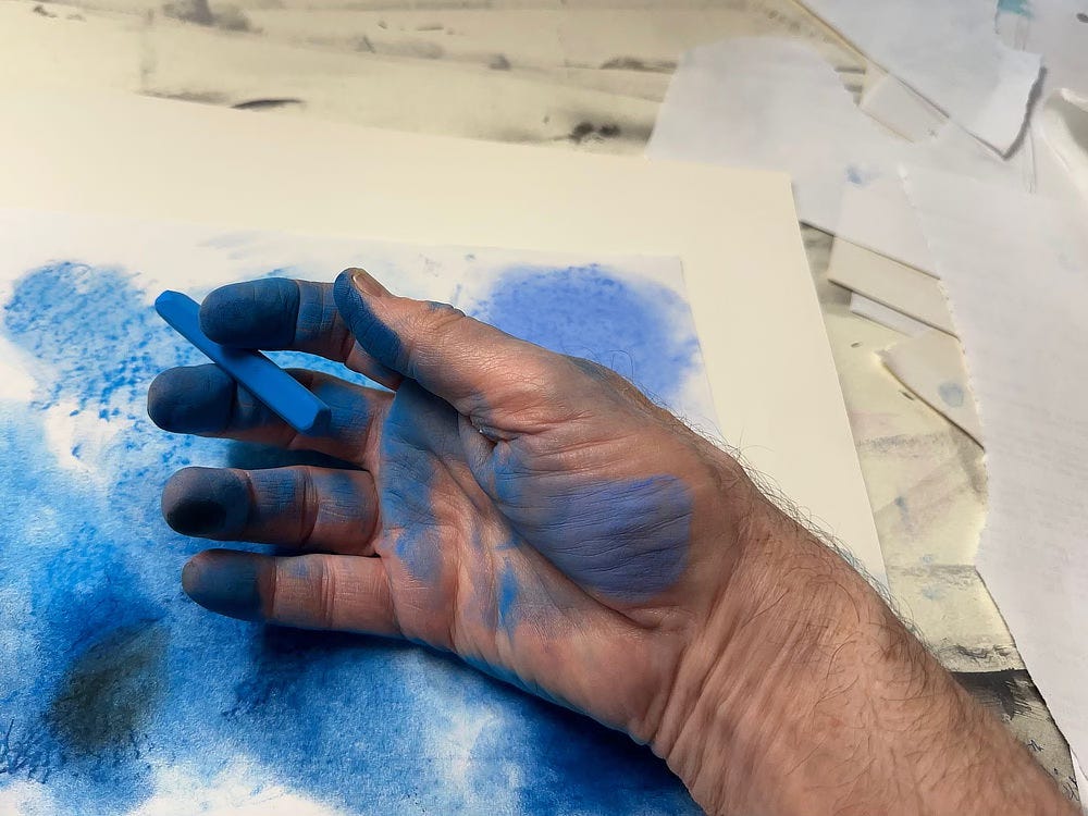 Photo of the artist's hand with a pastel stick between index and middle finger. His fingers are coated in blue and his palm is also marked. His hand rests on a stack of papers shaded blue.