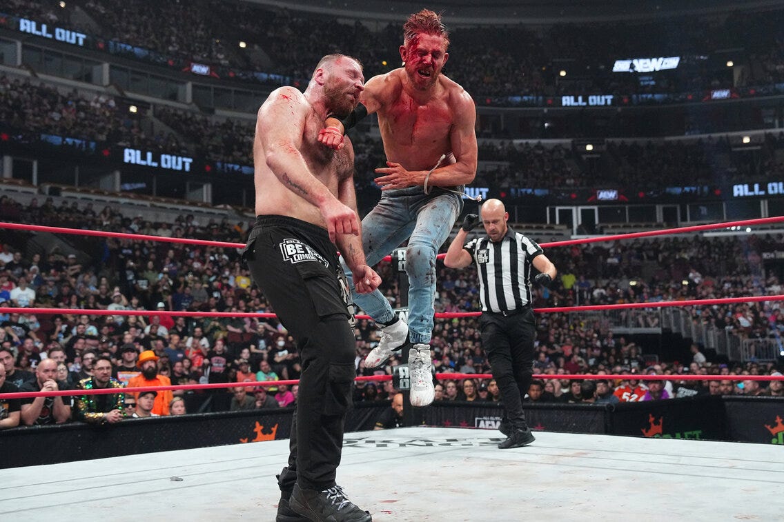Orange Cassidy, a white man in jeans, tussled blonde hair, and a bloodied face, leaping and delivering a punch to another white man with a crewcut, Jon Moxley, in black pants.