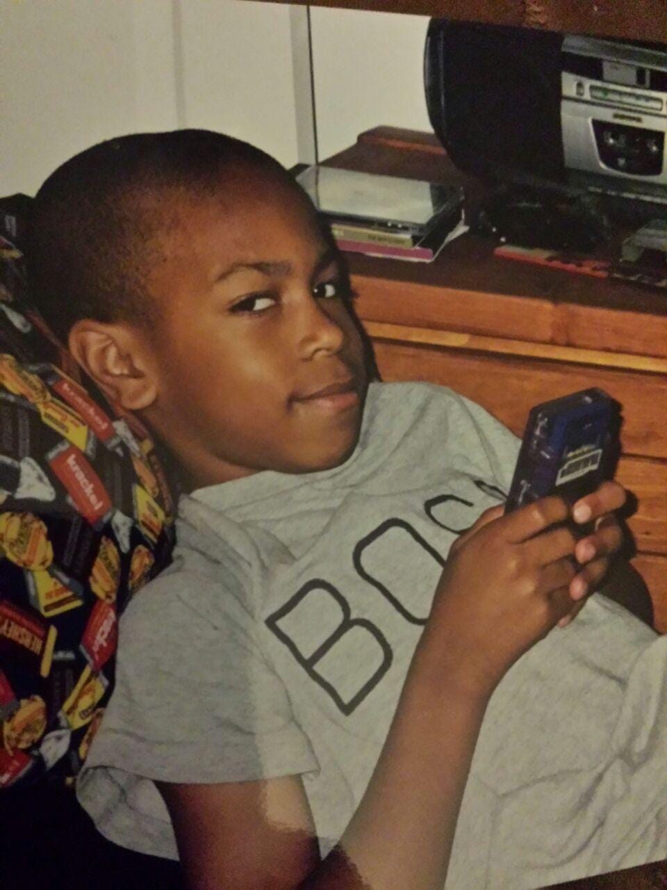young Ant, a skinny brown-skinned child, lays on a bed with a Gameboy in hand and a slight side eye to the camera. 