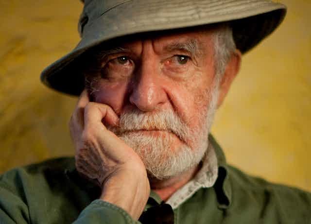 An elderly man with a grey beard and a khaki sun hat looks pensive, a hand to his jaw and his eyes looking into the distance. An elderly man with a grey beard and a khaki sun hat looks pensive, a hand to his jaw and his eyes looking into the distance.